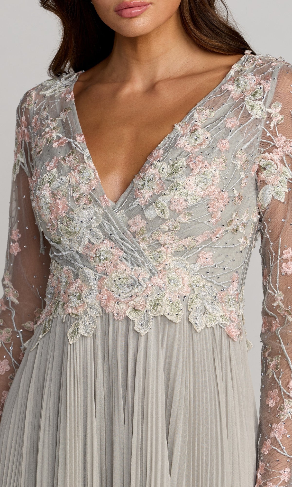 A woman wears the Mac Duggal 21332 long formal dress, featuring light gray chiffon with long sleeves, a deep V-neckline, and pink and white floral embroidery throughout its flowing A-line skirt.